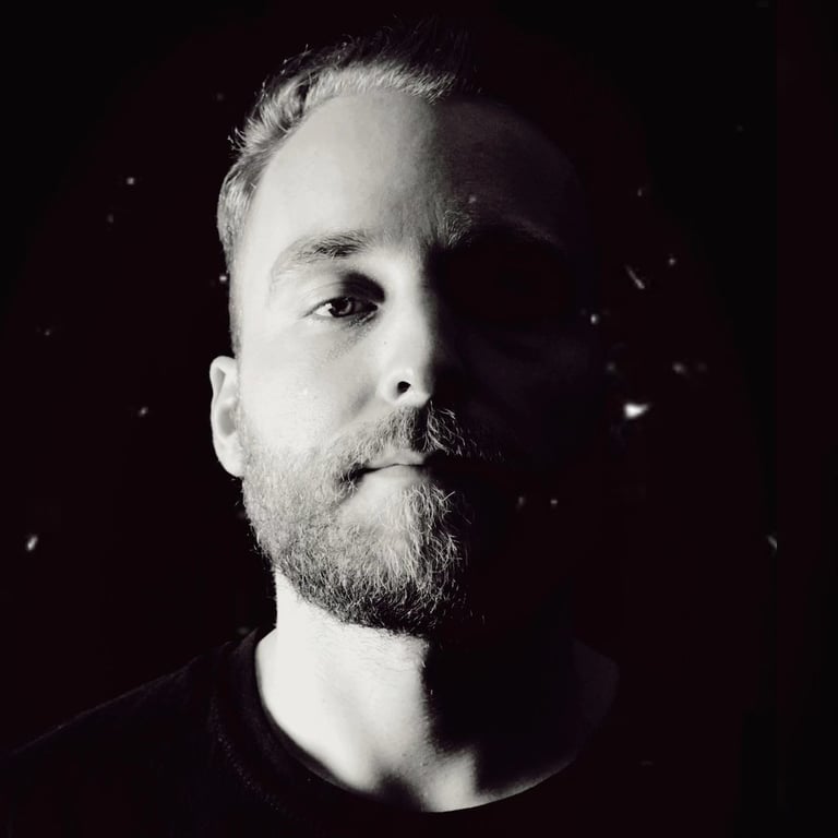 Black and white portrait of a man with a full beard, facing left against a dark background with subtle particle effects